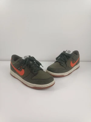 Nike Dunk Low SE Next Nature (GS) "Toasty Sequoia. Olivegrön. EUR 39 - De Nike Dunk Low SE Next Nature "Toasty Sequoia" (GS)-sneakers som visas är identifierade med stilkoden DC9561-300 och ingår i Nikes hållbarhetsinitiativ då de innehåller minst 20% återvunnet material räknat i vikt. Färgkombinationen beskrivs officiellt som Sequoia/Orange/Medium Olive/Sail, vilket ger skon en karaktärsfull look med en quiltad ovandel i mörk olivgrön textur, kompletterad med överdrag i mock. En framträdande orange Swoosh-logotyp i läder bryter av mot det gröna, och designen avslu