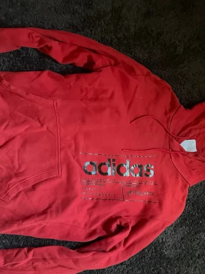Röd hoodie från Adidas med tryck - Snygg röd hoodie från Adidas med stor silverfärgad logga och text på bröstet. Klassisk känguruficka framtill och justerbar huva med dragsko. Tillverkad i mjukt bomullsmaterial som är skönt mot huden. Perfekt för en avslappnad streetwear-look.