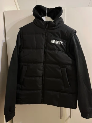 Svart hybridjacka från Hoodrich - Säljer en svart hybridjacka från Hoodrich i storlek XL för ungdomar. Jackan har huva, stora fickor och dragkedja framtill. Tydlig Hoodrich-logga på bröstet och ryggen. Perfekt för dig som vill ha en streetstyle-look.