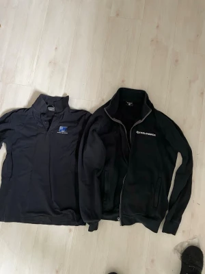 Svart fleecejacka & pikétröja - Två plagg: en svart fleecejacka med dragkedja och fickor från Gothblommor samt en svart pikétröja med krage och tryckt logga på bröstet. Fleecejackan har mjukt innerfoder och hög krage. Perfekt för dig som gillar enkel och funktionell stilsamt en Teddy fodrad väst finns små färgfläckar på  två av plaggen därav priset 