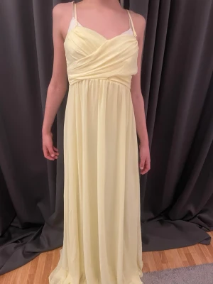 Ljusgul långklänning med smala axelband - Elegant ljusgul långklänning med smala axelband och omlottdetalj över bysten. Klänningen har ett luftigt fall och markerad midja, perfekt för sommarens tillställningar. Tillverkad i ett lätt och skönt polyestertyg. Man kan även ta av banden. .