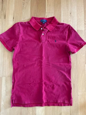 Röd pikétröja från Polo Ralph Lauren - Klassisk röd pikétröja från Polo Ralph Lauren med korta ärmar, krage och två knappar vid halsen. Liten blå logga broderad på bröstet. Tillverkad i mjuk bomull som känns skön mot huden. Perfekt för en avslappnad men ändå stilren look. De är en bran XL men sitter som en S