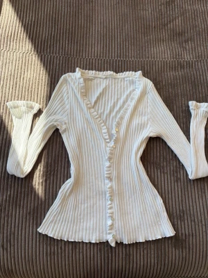 Vit ribbad kofta med volangkant från Odd Molly - Säljer en söt vit ribbad kofta med volangkant längs knäppningen och små pärlemorknappar. Koftan har långa ärmar och figurnära passform, perfekt för att lyfta vilken outfit som helst. Materialet är mjukt och stretchigt.Superbra skick!,skickar inom 1-2 dagar