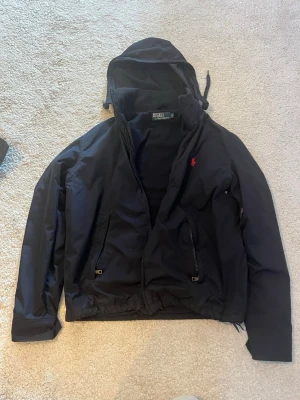 Svart vindjacka från Polo Ralph Lauren - Snygg svart vindjacka från Polo Ralph Lauren med huva och dragkedja framtill. Jackan har två framfickor med dragkedja, knappdetaljer vid ärmsluten och den klassiska röda loggan broderad på bröstet. Materialet är tunt och vindtätt, perfekt för blåsiga dagar.