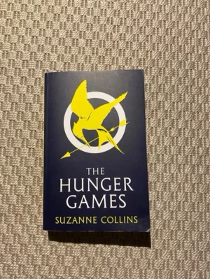 The Hunger Games pocketbok - Säljer boken 'The Hunger Games' av Suzanne Collins. Omslaget är mörkblått med en gul symbol av en fågel och pil. Baksidan är gul med svart text. Perfekt för dig som gillar dystopiska äventyr och spännande berättelser.