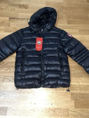 Svart dunjacka från Canada Goose - Snygg svart dunjacka från Canada Goose med huva och två stora dragkedjefickor framtill. Jackan är strl S den är ny aldrig använd inga skador eller något sånt såklart den är jätte bra kvalle och tar byten och pris kan diskuteras den funkar både för vår och höst tycker jag 