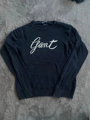 Mörkblå stickad tröja från Gant - Snygg mörkblå stickad tröja från Gant med vit broderad logga framtill. Tröjan har rund halsringning, långa ärmar och ribbade muddar vid ärmslut och nederkant. Perfekt för en stilren och avslappnad look. Hör av dig vid intresse eller om du har några övriga funderingar 🙌