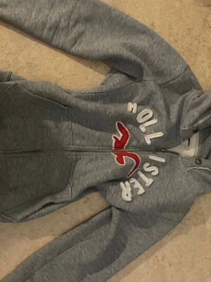 Grå Hollister hoodie med dragkedja - Snygg grå hoodie från Hollister med dragkedja framtill och stor huva. Framsidan har vit brodyrtext och den klassiska röda loggan. Tillverkad i mjukt sweatshirtmaterial och har två fickor fram. Perfekt för en avslappnad och trendig look.