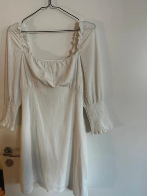 Vit kortklänning med snörning - Söt vit kortklänning med diskreta ränder, vida trekvartsärmar med smock och volangkant. Klänningen har en djup rygg med snygg snörning och volangdetaljer vid urringningen. Perfekt för sommarens alla tillfällen. Endast använd på mitt utspring på studenten. Rök och djurfritt hem. Pruta gärna!🌸