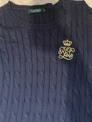 Mörkblå kabelstickad tröja Lauren - Snygg mörkblå kabelstickad tröja från Lauren Ralph Lauren med rund halsringning och ett broderat RL-emblem i guld på bröstet. Tröjan har långa ärmar och ribbade muddar. Perfekt för en klassisk och stilren look.