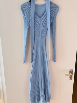Ljusblå ribbad långklänning - Supersnygg ljusblå långklänning med ribbad struktur och v-ringning. Klänningen har långa ärmar och ett matchande bälte i midjan för extra stilpoäng. Perfekt för dig som vill ha en trendig och bekväm look.