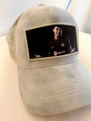 Velvet keps-Lamine Yamal - Säljer en ljusgrå keps med mesh på sidorna och ett stort tryck framtill av Lamine Yamal i matchtröja med Spotify-logga. Kepsen har böjd skärm och justerbar baksida. Perfekt för dig som gillar fotboll eller för att vara stilig och vill sticka ut.
