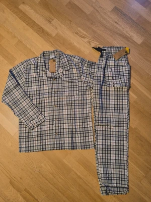 Rutiga pyjamas i bomull - Klassisk tvådelad pyjamas med långärmad skjorta och byxor i blåvitt rutigt mönster. Skjortan har knappar framtill och bröstficka, byxorna har resår i midjan. Tillverkad i mjuk bomull, perfekt för mysiga kvällar hemma.
