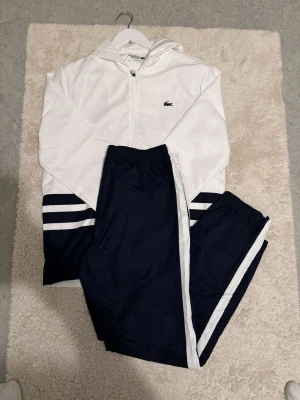 Vit och marinblå Lacoste loungewear - Snyggt set från Lacoste med vit zip-hoodie och marinblå byxor. Hoodien har dragkedja, huva och den klassiska Lacoste-loggan på bröstet. Byxorna är marinblå med vita ränder på sidorna och resår i midjan. Perfekt för chill dagar hemma.