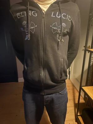 Svart hoodie från Bling Lucid  - Snygg svart hoodie från Bling Lucid i storlek XS men den passar S mest skulle jag säga. Dragkedja framtill, huva med snören och fickor på sidorna. Dekorerad med glittriga stenar som formar texten 'BLING LUCID' och två cupider på bröstet. Perfekt för dig som gillar streetwear och detaljer. Små fläckar som går bort i tvätten.