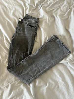 Grå raka jeans med slitning - Säljer ett par grå jeans med rak passform och diskret slitning vid ena låret. Klassisk femficksmodell med knapp och dragkedja framtill. Jeansen har en tvättad look och är tillverkade i bomullsmix för skön känsla.