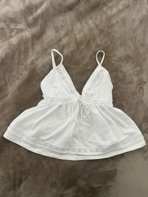 Vit BNWT babydoll linnetopp från Gina Tricot - Säljer en helt oanvända ny med lappen kvar söt vit babydoll linnetopp från Gina Tricot med smala axelband, v-ringning och dekorativa spetsdetaljer. Toppen har knyt i midjan och lätt utställd passform, perfekt för varma dagar.