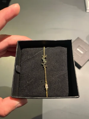 Guldigt armband från Saint Laurent - Snyggt och stilrent armband i guldton från Saint Laurent med den ikoniska YSL-loggan framtill. Kedjan är tunn och justerbar med ett litet hjärta och en bricka med märkets namn på baksidan. Perfekt accessoar för dig som gillar exklusiv och trendig design.