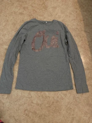 Grå långärmad topp med glittertext - Snygg grå långärmad topp med rosa glittertexten 'Oui' på bröstet. Tillverkad i mjukt bomullsliknande material och har en avslappnad passform. Perfekt för dig som gillar enkla men ändå trendiga plagg med en touch av glitter.