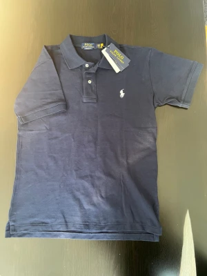 Mörkblå piké från Polo Ralph Lauren - Snygg mörkblå pikétröja från Polo Ralph Lauren med klassisk krage och korta ärmar. Tröjan har den ikoniska loggan broderad på bröstet och är tillverkad i mjuk bomull för skön känsla. Perfekt till jeans eller shorts för en clean look.