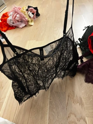 Svart spetsbody med tunna band - Säljer en svart body i transparent spets med tunna, justerbara axelband. Bodyn har en snygg meshdetalj och vacker blommig spets som ger en elegant look. Aldrig använt den, den är tvättad. 