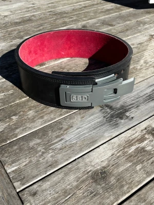 SBD svart träningsbälte - Säljer ett svart träningsbälte från SBD med röd insida och kraftigt metallspänne. Bältet är gjort i rejält skinn och har flera hål för justering. Perfekt för dig som vill ha extra stöd vid styrketräning. Bältet har tecken på användning men det är inget som påverkar funktionen. Storleken är M/L