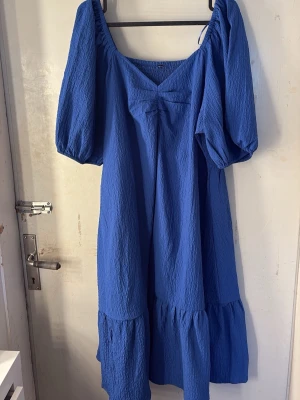 Blå volangklänning med puffärm - Säljer en blå klänning med volangkant och puffärmar. Klänningen har en lös passform, fyrkantig ringning och är tillverkad i ett strukturerat tyg som känns luftigt. Perfekt för dig som gillar en avslappnad men trendig stil. 
