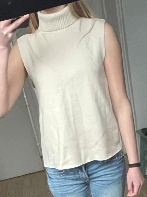 Beige ärmlös polotopp - Stilren beige polotopp utan ärmar med ribbad hög krage. Toppen har en lös passform och är tillverkad i ett mjukt stickat material som känns skönt mot huden. Perfekt att styla med jeans eller kjol för en clean look.