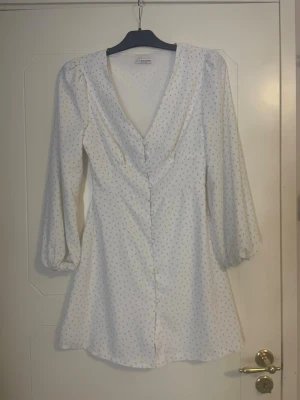 Vit blommig klänning Nelly x Glamorous - Söt vit kortklänning från Nelly x Glamorous med små blommiga detaljer i pastellfärger. Klänningen har v-ringning, knappar framtill och långa ärmar med lätt puff. Perfekt för sommarens alla tillfällen! Ett knapp snöre har gått sönder men de går enkelt att sy fast💕