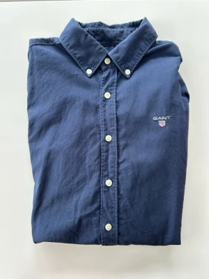 Mörkblå skjorta från Gant - Klassisk mörkblå skjorta från Gant med vit knappstängning och broderad logga på bröstet. Skjortan har lång ärm, normal passform och är tillverkad i mjuk bomull. Perfekt till jeans eller chinos för en clean och stilren look. Storlek 158-164 cm 