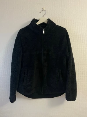 Svart teddyjacka med dragkedja - Mysig svart teddyjacka med hög krage och hel dragkedja framtill. Jackan har två sidofickor och är gjord i fluffigt, mjukt material som håller dig varm. Perfekt för lager på lager och en chill look.