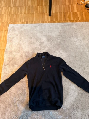 Polo Ralph lauren quarter zip - Snygg marinblå tröja med halv dragkedja från Polo Ralph Lauren. Klassisk röd logga broderad på bröstet och ribbade muddar vid ärmslut och nederkant. Perfekt till jeans eller chinos för en clean och stilren look.