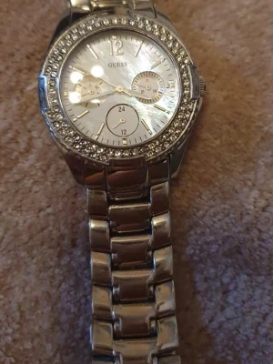 Silverfärgad klocka från Guess - Stilren klocka från Guess i silverfärg med länkarmband i rostfritt stål. Urtavlan är dekorerad med gnistrande stenar runt boetten och har flera små urtavlor för extra funktioner. Perfekt accessoar för dig som gillar bling och klassisk design.