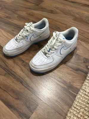 Vita Nike Air Force 1 med blå detaljer - Säljer ett par klassiska vita Nike Air Force 1 sneakers med blå detaljer på swoosh och sula. Skorna har perforerad tå, vita skosnören och låg profil. Perfekta för dig som gillar stilrena och tidlösa sneakers. Lite smutsiga men inga repor och dom är nästan helt nya. Storlek 42,5.  Skriv frågor!