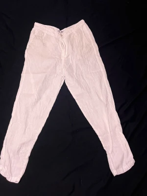 Vita linnebyxor från Zara - Säljer ett par vita linnebyxor från Zara med avslappnad passform och raka ben. Byxorna har dragkedja och knapp framtill och är tillverkade i ett lätt och luftigt linnetyg, perfekta för varma dagar.