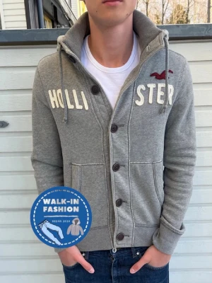 Grå kofta med knappar från Hollister - Snygg grå kofta från Hollister med stora knappar framtill, broderad logga och text på bröstet. Tröjan är i 9/10 skick med passform för 178-179 men passar snyggt för uppemot 185