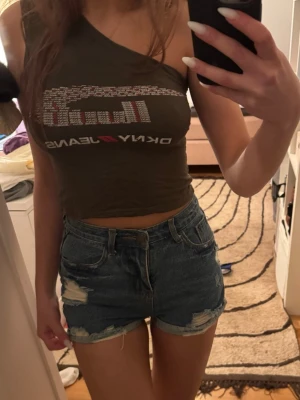 Blå denimshorts med slitningar - Säljer ett par blå denimshorts med råa slitningar och fransiga detaljer. Klassisk femficksmodell med hög midja och knappgylf. Perfekta till sommaren för en avslappnad och trendig look.