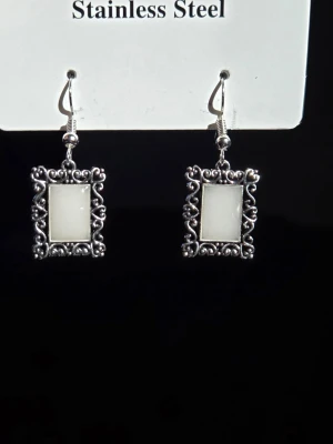 Rektangulära glow örhängen i rostfritt stål - Lyser lila!  - Snygga örhängen i rostfritt stål med rektangulär form och dekorativ, antik-inspirerad ram. Mittdelen är vit i dagsljus och lyser lila i mörker. Effekten sköter sig själv med hjälp av dagsljuset utifrån när du bär dom, solljus fungerar också. Krokarna är i rostfritt stål och örhängena tål vatten. Hängena lyser i flera timmar av normal användning. Lång livslängd. Perfekta för dig som gillar unika accessoarer med vintagekänsla. Handgjort, begränsat lager.