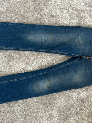 Blå jeans från Nudie Jeans - Säljer ett par klassiska blå jeans från Nudie Jeans med snygga slitningar och kontrastsömmar. Jeansen har raka ben och bakfickor med den ikoniska Nudie-bågen i orange. Tillverkade i denim med en cool tvättad look. Str 30/32 