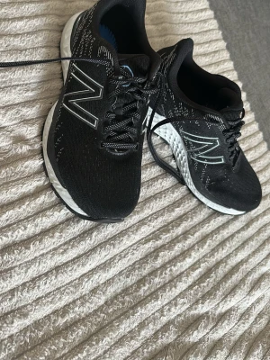 Svarta New Balance sneakers  - Svarta New Balance sneakers med vit sula och reflekterande N-logga på sidorna. Ovandelen är i ventilerande mesh och skorna har klassisk snörning. Sulan är dämpad för extra komfort och insidan är blå. Perfekt för dig som gillar sportig stil.
