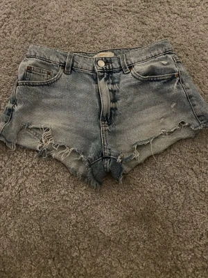 Gråblå jeansshorts från Gina Tricot - Säljer ett par gråblå jeansshorts från Gina Tricot med råa, fransiga kanter och slitna detaljer. Shortsen har fem fickor, bälteshällor och knappgylf. Perfekta för sommaren och ger en avslappnad vibe.