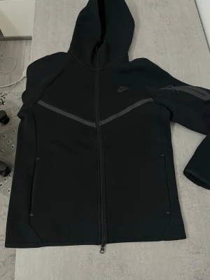 Svart Nike tech med nya modellen  - Inga hål eller nånting har haft på mig den 5 eller 6 gånger den är plain black inte sol bränd och färgen är hel svart köpt den för 1500 ny från Nike hem sidan 