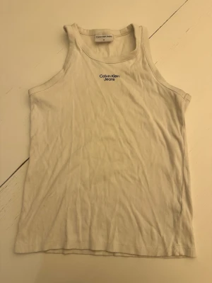 Vit ribbad linnetopp Calvin Klein Jeans - Säljer en vit ribbad linnetopp från Calvin Klein Jeans med broderad logga framtill. Toppen har en klassisk, ärmlös design och är tillverkad i mjuk bomull som känns skön mot huden. Perfekt att styla med jeans eller kjol för en clean look.