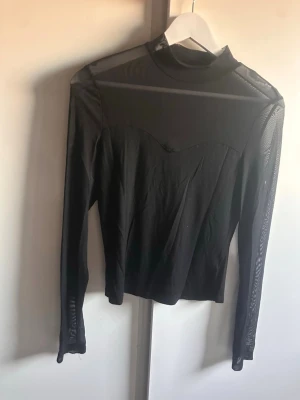 Svart mesh polotopp med långa ärmar - Trendig svart polotopp i mesh med långa ärmar och transparenta partier. Toppen har en figurnära passform 