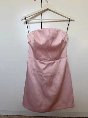 Ljusrosa axelbandslös kort klänning - Söt ljusrosa axelbandslös klänning med enkel och stilren design. Klänningen har en figurnära passform och är sydd i ett slätt material som ger en elegant look. Perfekt för dig som vill ha en feminin och klassisk stil.