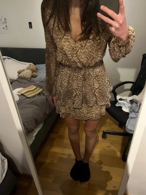 Brun mönstrad kortklänning - Säljer en snygg kortklänning i beige och bruna toner med ett diskret ormmönster. Klänningen har v-ringning, långa ärmar och resår i midjan