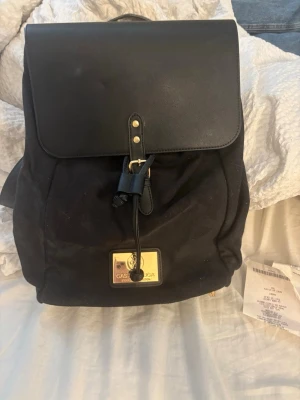 Svart ryggsäck från Gant Gaston Luga - Snygg svart ryggsäck från Gaston Luga med lock i läderimitation och dragskostängning. Väskan har guldfärgade detaljer och en tydlig logga framtill. Materialet är en mix av tyg och konstläder, och insidan har en röd detalj. Perfekt för dig som vill ha en stilren och praktisk ryggsäck.