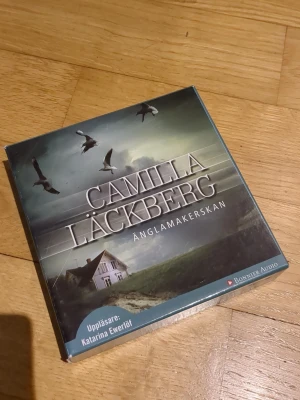 Änglamakerskan av Camilla Läckberg - Ljudbok på CD, 'Änglamakerskan' av Camilla Läckberg, utgiven av Bonnier Audio. Omslaget är mörkt med en bild på ett hus, fåglar och dramatisk himmel. Uppläsare är Katarina Ewerlöf. Perfekt för dig som gillar spänning och deckare.