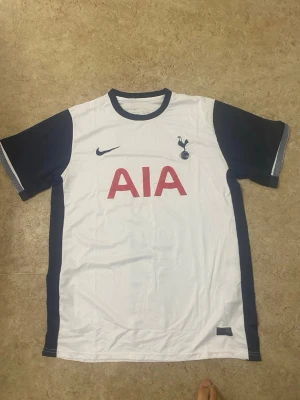 Tottenham Hotspur matchtröja Nike - Snygg Tottenham Hotspur matchtröja från Nike i vitt med mörkblå ärmar och detaljer. Stor röd AIA-logga på bröstet och klubbmärke på vänster sida. Tillverkad i lätt och ventilerande material, perfekt för fotboll eller träning.