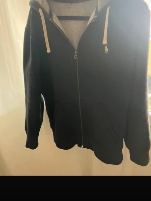 Svart zip hoodie från Polo Ralph Lauren - Säljer en svart hoodie med dragkedja från Polo Ralph Lauren. Hoodien har vit broderad logga på bröstet, vita snören i huvan och två fickor framtill. Insidan är mjuk och materialet känns som bomullsblandning. Perfekt för en chill och stilren look.